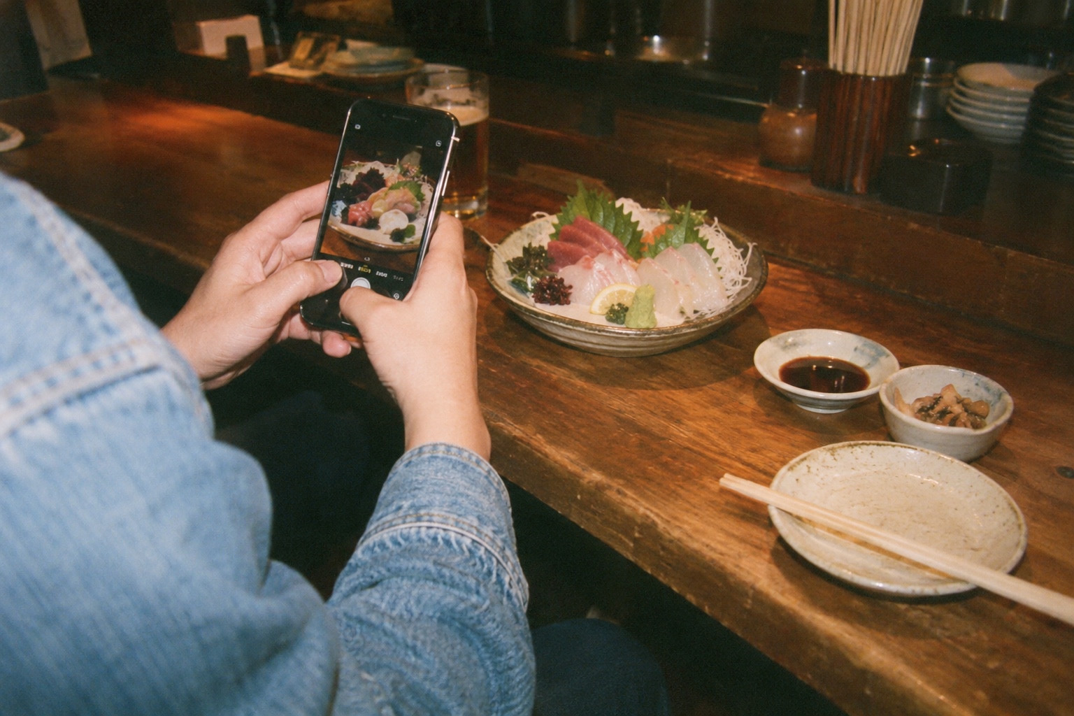 木のテーブルに並ぶ刺身をスマートフォンで撮影するイメージ